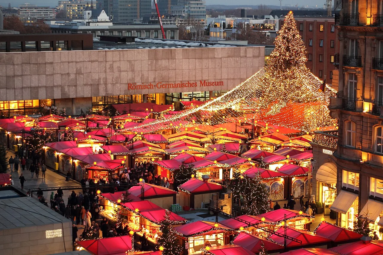 Glowing Christmas market stalls and a lit Christmas tree beside Cologne Cathedral.